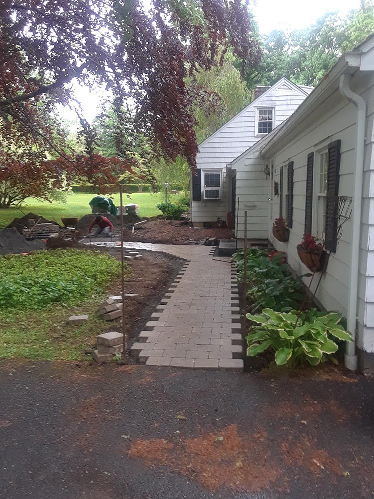 Narrow brick walkway beside a white house with hanging plants, leading to a green yard and garden area.