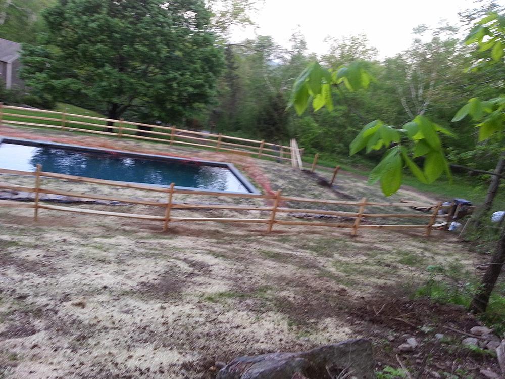 Outdoor swimming pool surrounded by a wooden fence and trees on a gravelly hillside.