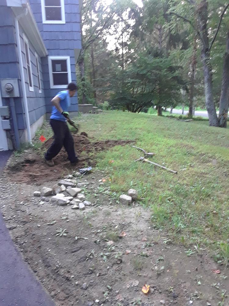 Person shoveling dirt and rocks beside a house in a grassy yard.