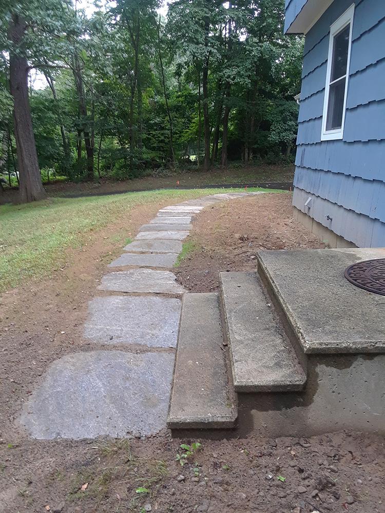 Concrete stepping stones beside blue house leading into a grassy yard near trees