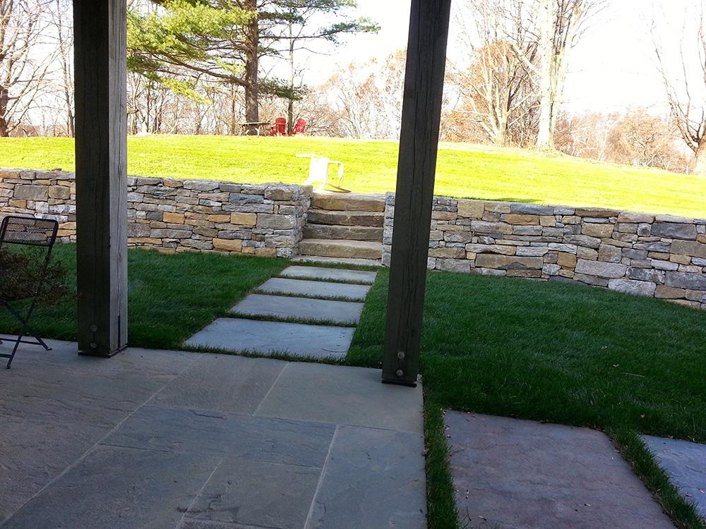 Covered patio opening to a stone path, retaining wall, and grassy yard in bright daylight