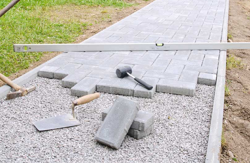Broken paving stones and tools scattered on a gravel path beside a partially repaired walkway.