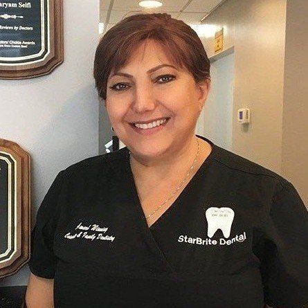 A woman in a black scrub top is smiling in a dental office.