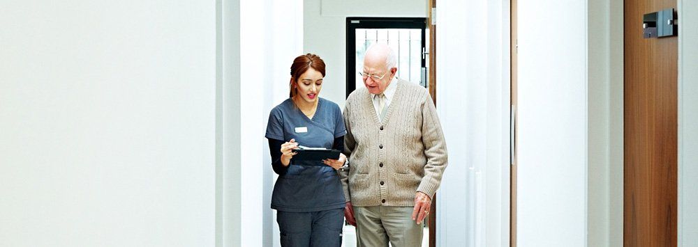 A nurse is helping an elderly man walk down a hallway.