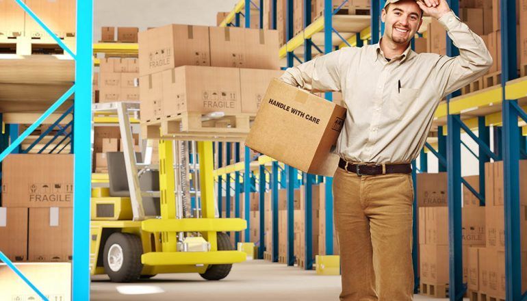Warehouse worker holding box, smiles. Forklift with boxes on pallets nearby. Shelves in background.