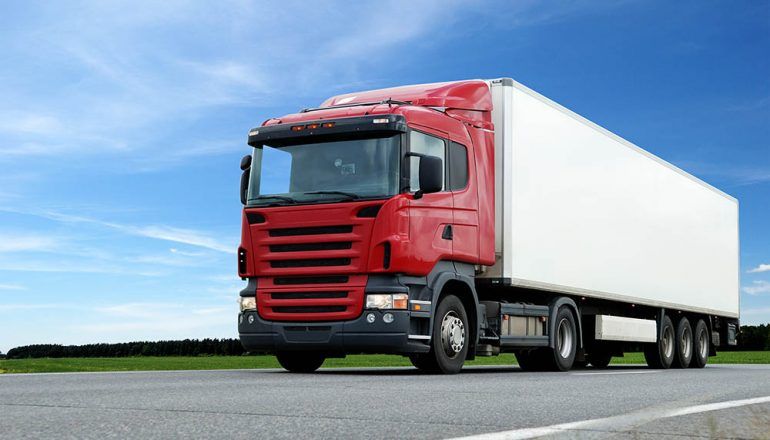 Red semi-truck with a white trailer driving on a road under a blue sky.