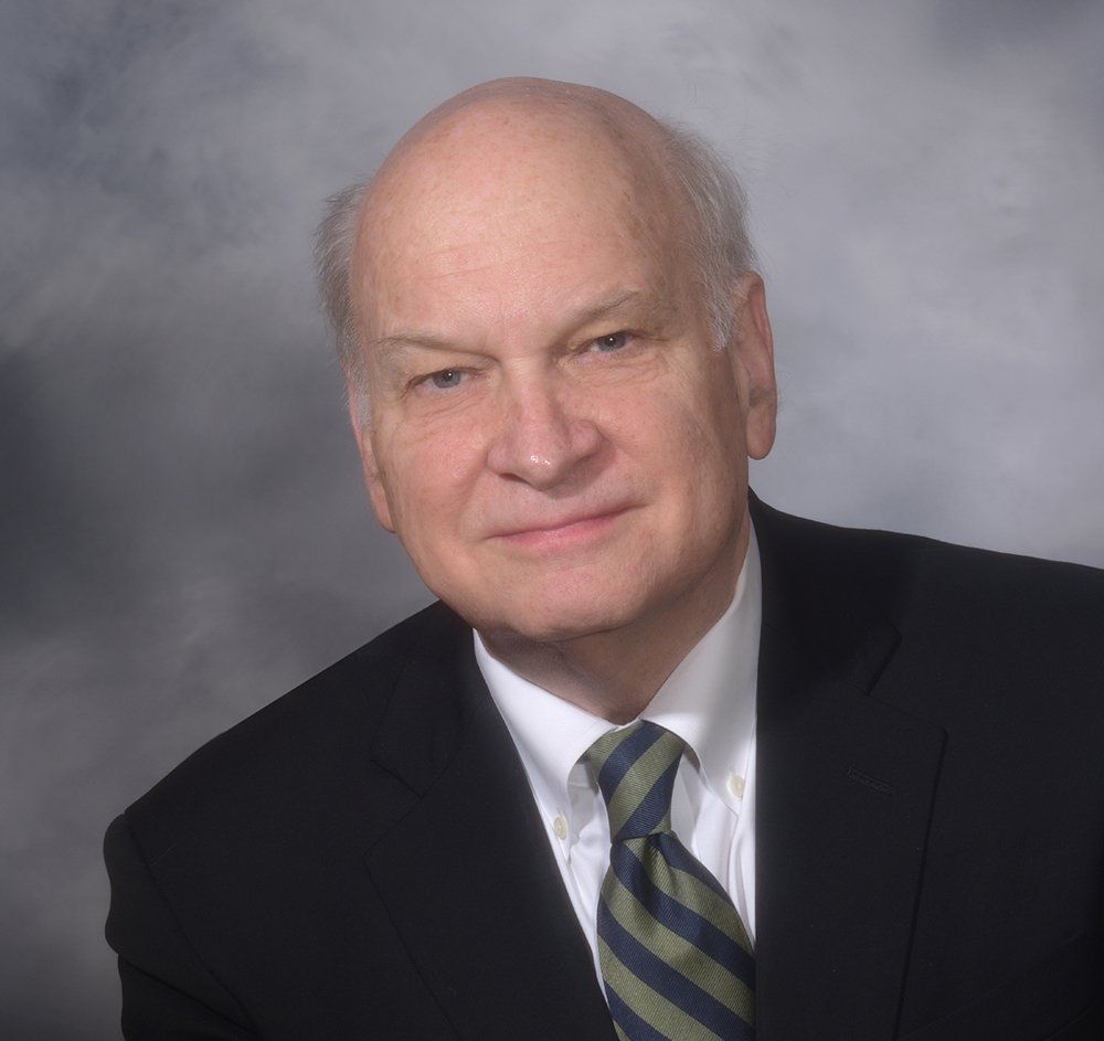 Portrait of a man in a black suit, white shirt, and striped tie against a gray studio background