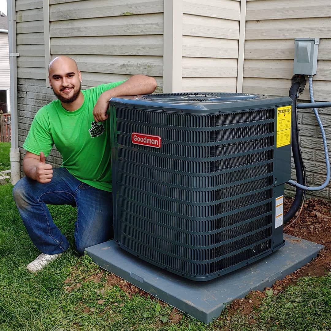Man in green shirt kneels next to a Goodman air conditioner, giving a thumbs up.
