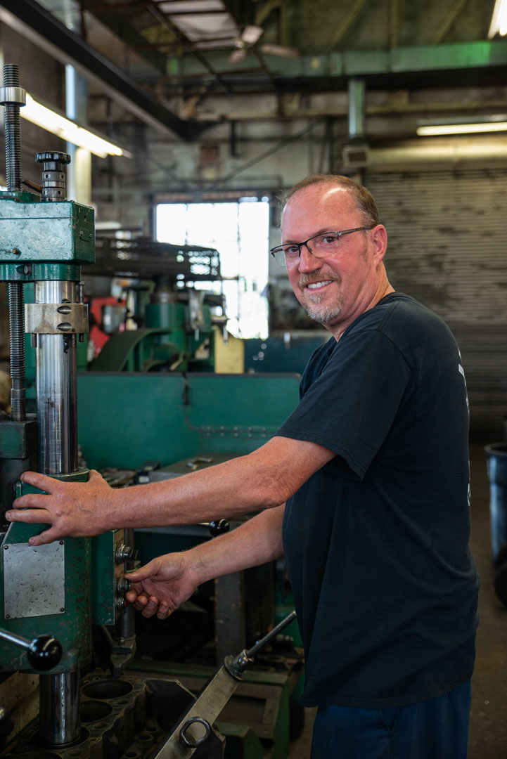 Man in glasses smiles, operating a green industrial machine in a workshop.