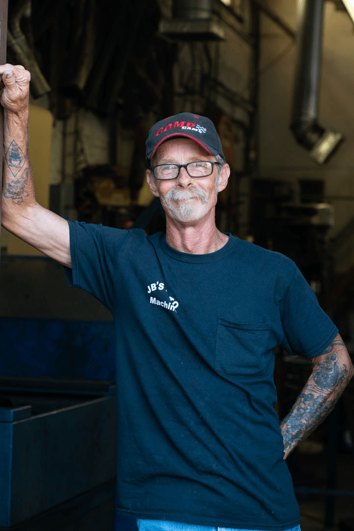 Man with tattoos and glasses leans against equipment in a workshop, wearing a hat and dark t-shirt.