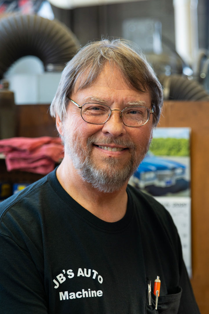 Man in black shirt, glasses, smiling; inside auto shop with equipment and calendar visible.