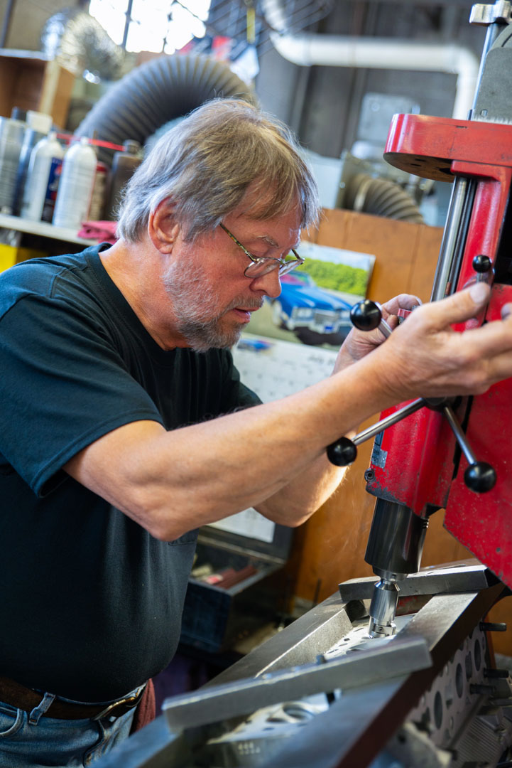 Man using a drill press in a workshop. Red machine, focused expression, gray hair, black shirt.