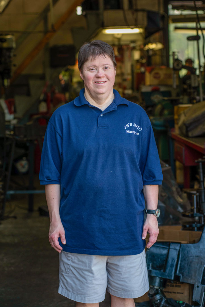 Person in a blue polo shirt and gray shorts stands in a workshop, smiling.