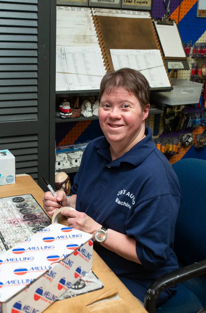 A smiling person with Down syndrome in a blue shirt, writing at a desk in a store.