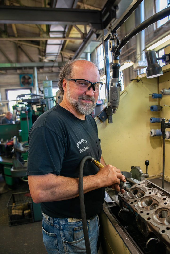 Man in glasses working on engine head in a workshop. Smiling, holding tool.