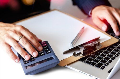Hands using calculator and laptop, documents on clipboard, working at a desk.