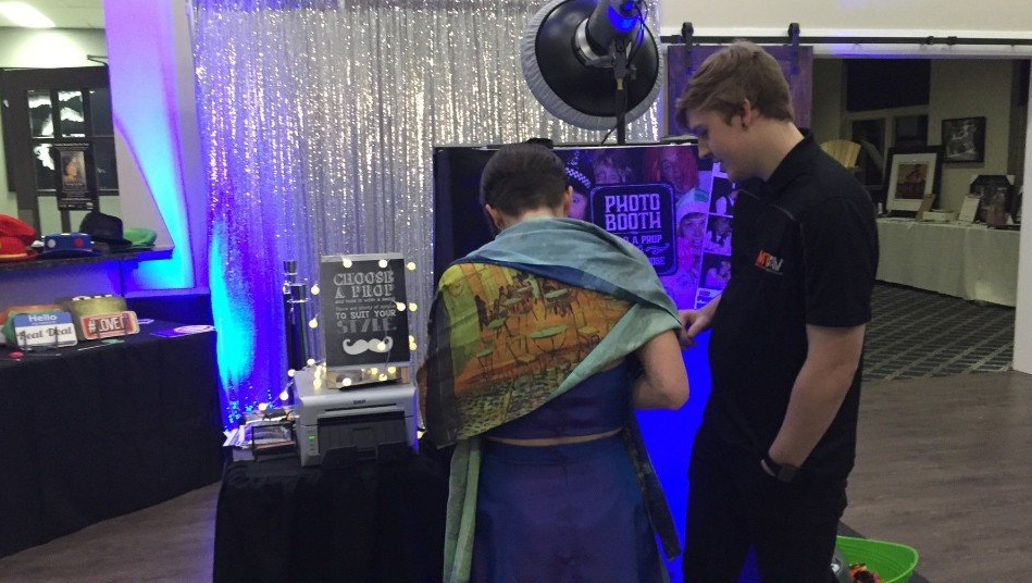 Person operating a photo booth. One person looks at the screen, and another person looks on. Silvery backdrop with blue lights.