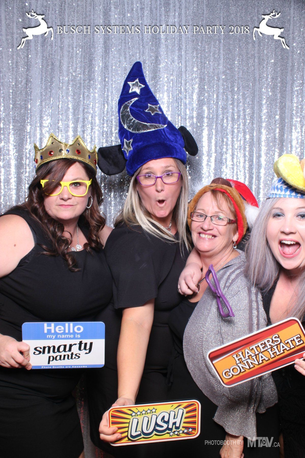 Four people posing for a photo booth with props against a silver backdrop.