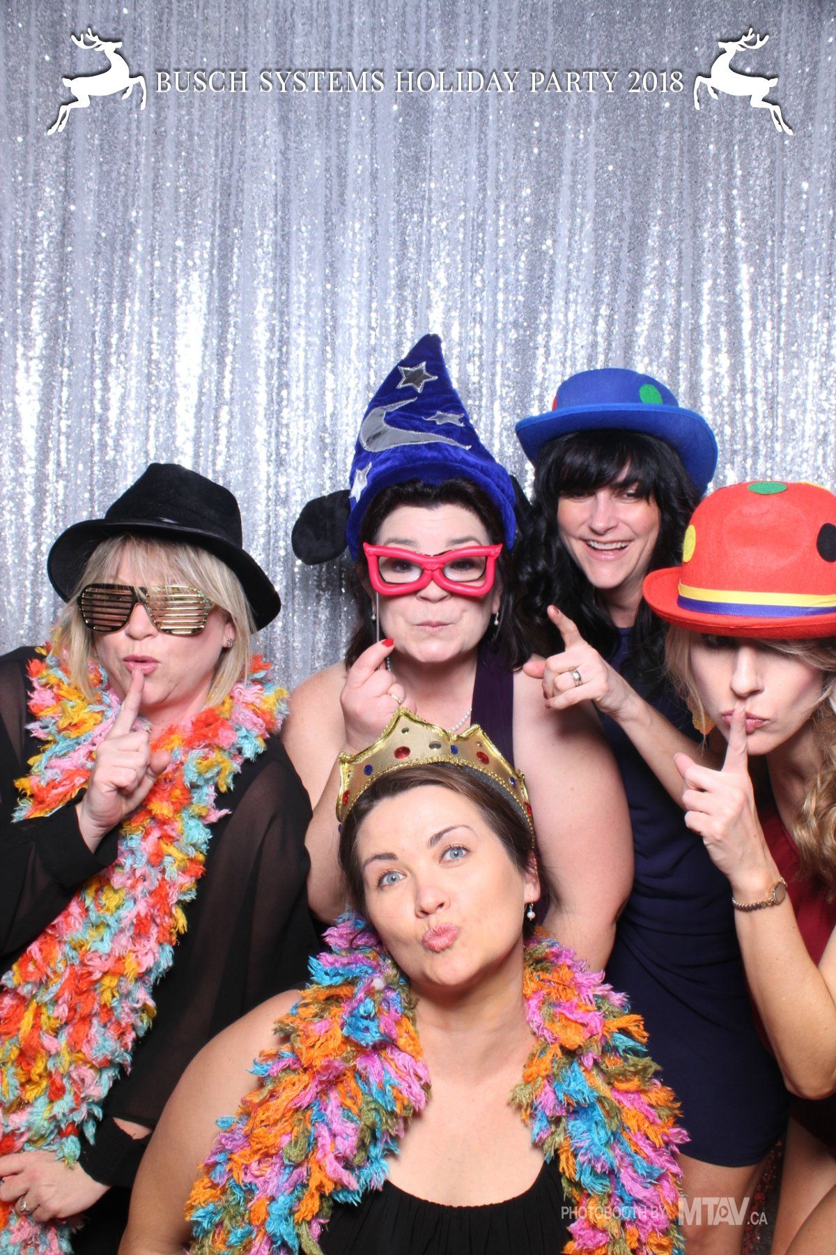 Five women pose in a photo booth with props, making silly faces and gestures. A shiny silver backdrop is behind them.