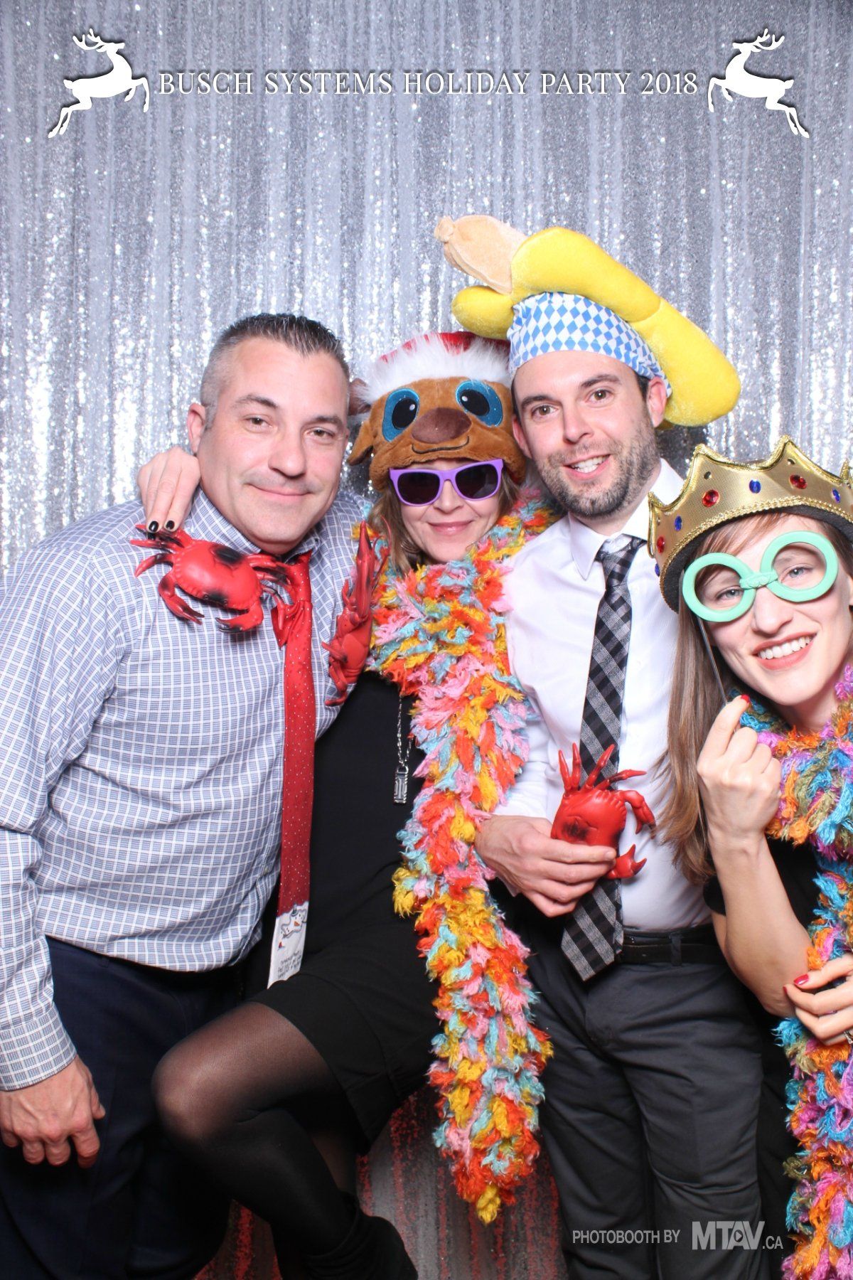 Four people in a photo booth wearing festive accessories against a silver sequin backdrop.