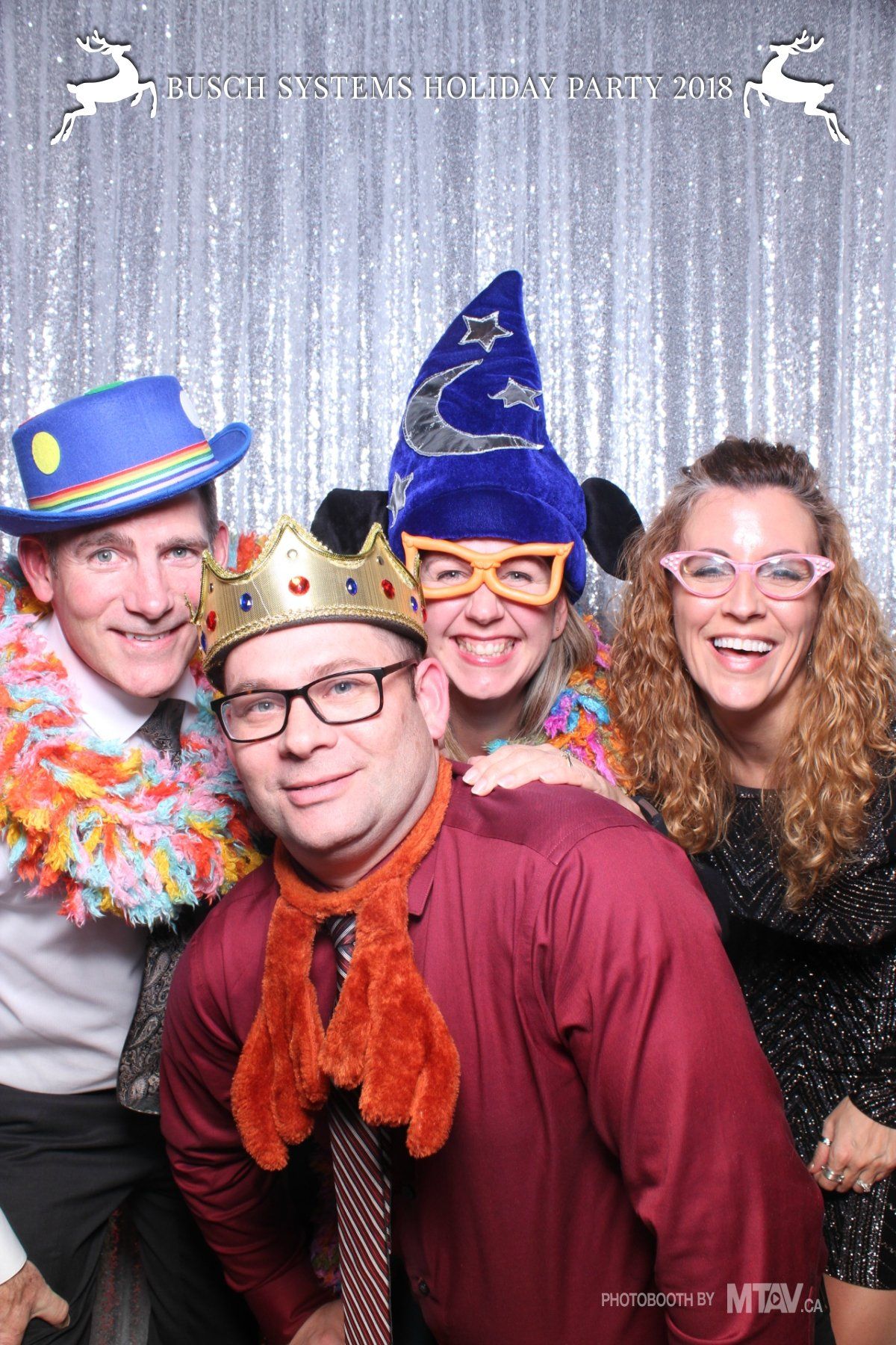 Four people in festive attire posing for a photo booth. Silver backdrop. Smiles, hats, and glasses.