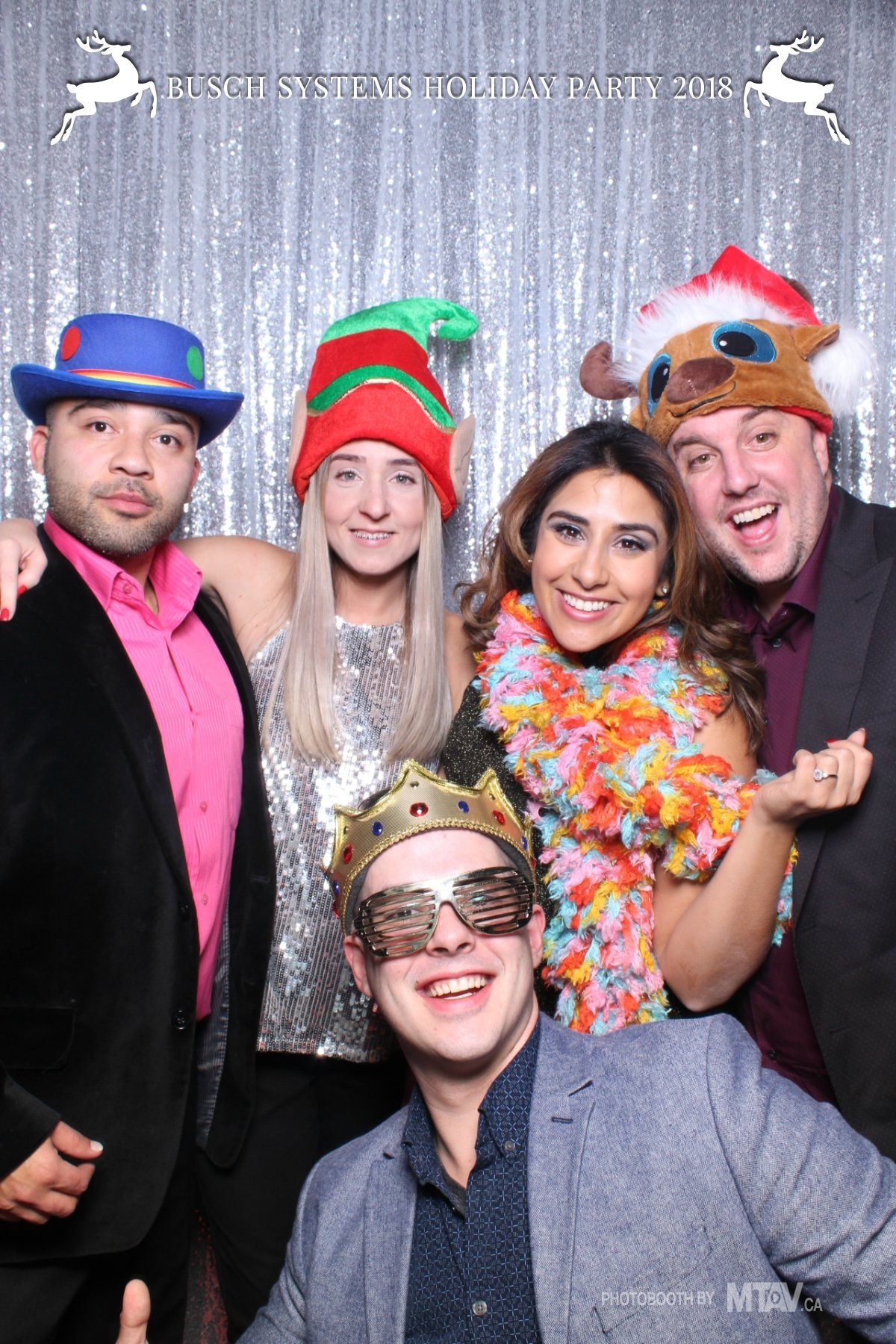 Five people in a photo booth, wearing festive hats and accessories, smiling in front of a silver backdrop.