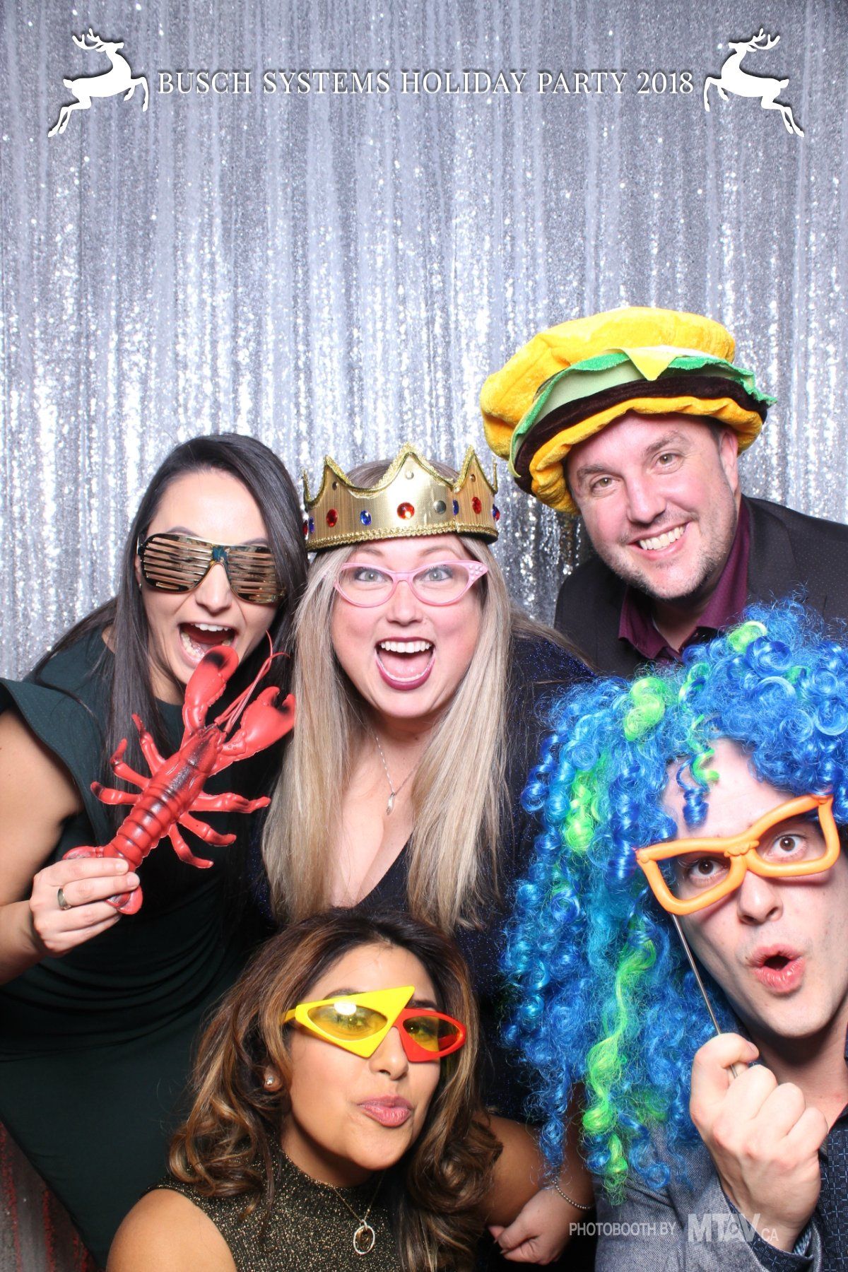 People at a holiday party posing with props like a lobster claw, crown, hamburger hat, and wigs.