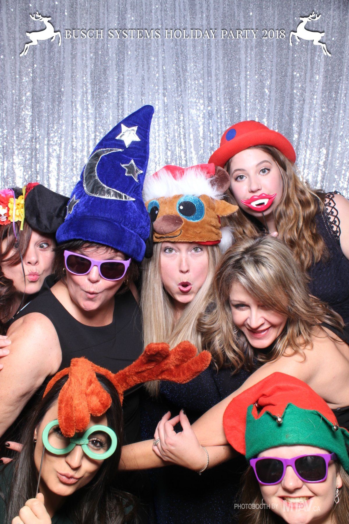 Group of women in festive hats, posing for a photo booth with silly faces and glasses in front of a sparkly backdrop.