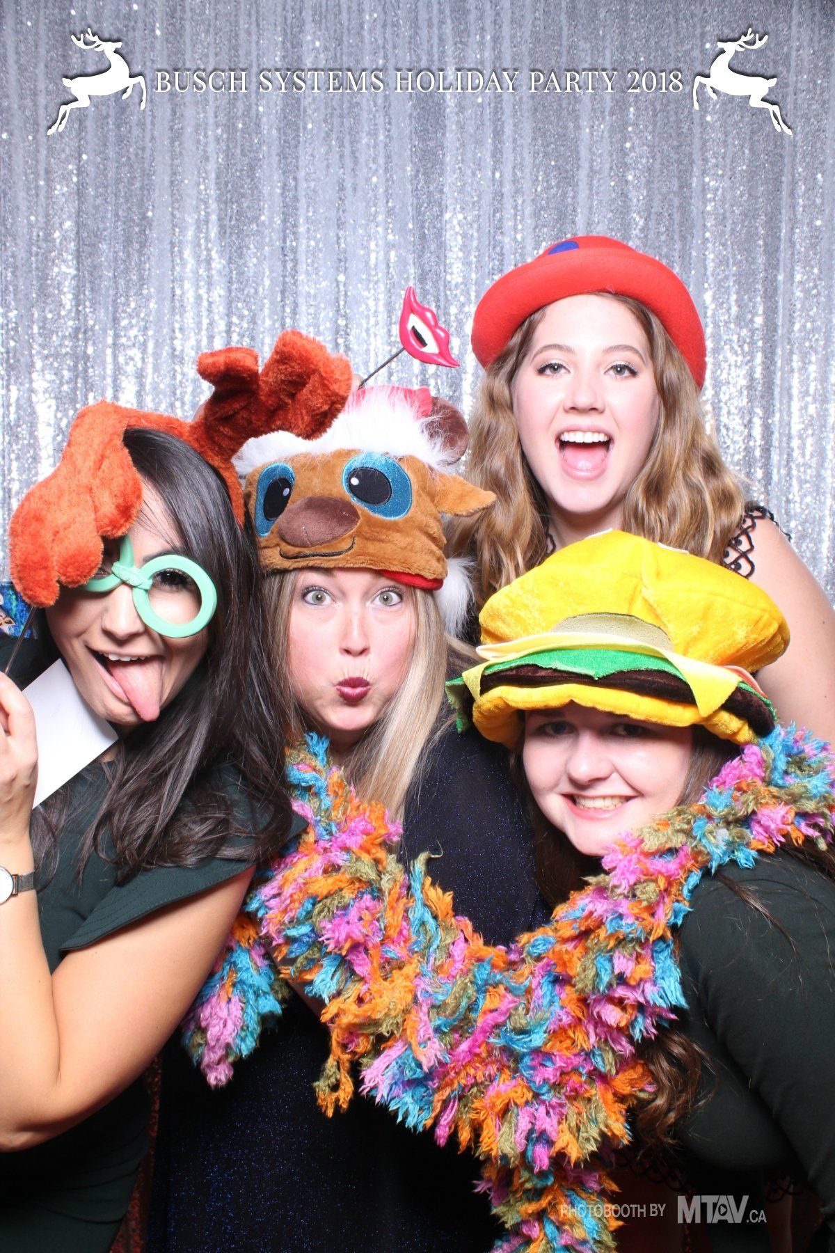 Four people posing for a photo, wearing festive hats and boas, making silly faces. Silver sparkly backdrop.