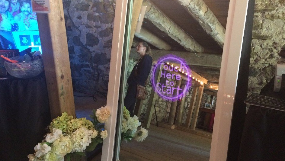 Photo booth mirror at an event with flowers.  A person reflected in the mirror, and a purple graphic overlay reads 