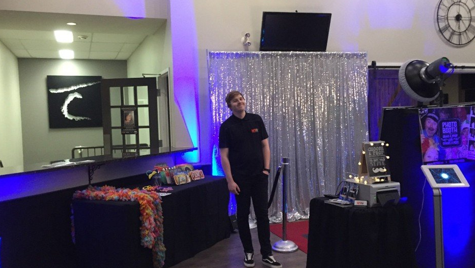 Man stands near a photo booth with a silver backdrop. The booth and table are black. Purple and blue lighting.