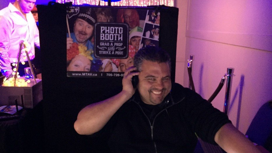 DJ wearing headphones smiles, stands near photo booth with purple lighting.