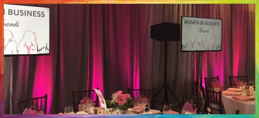 A women in business event with signs, tables, and flowers. Pink and gray drapery backdrop.