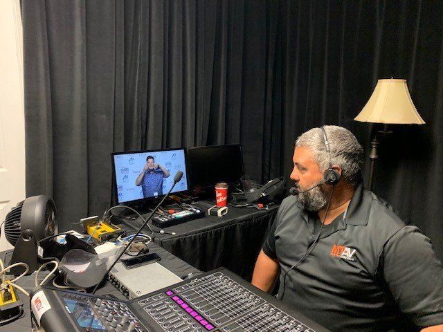 Man with headset at audio mixing board; another person on screen. Black backdrop.