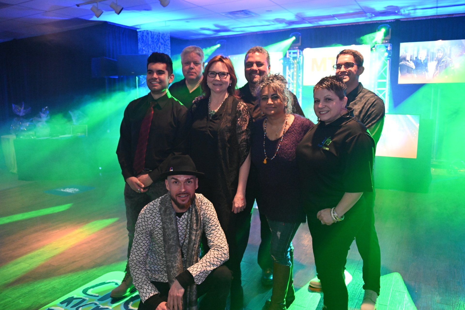 Group of nine people posing on a stage with colorful lighting and a fog effect.