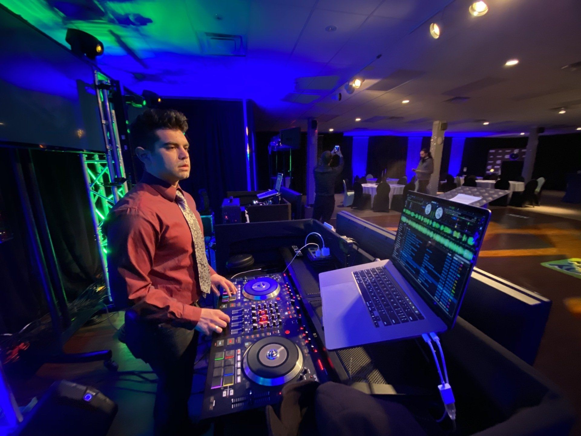 DJ in red shirt and tie at a party, using a mixer and laptop with blue and green lighting.