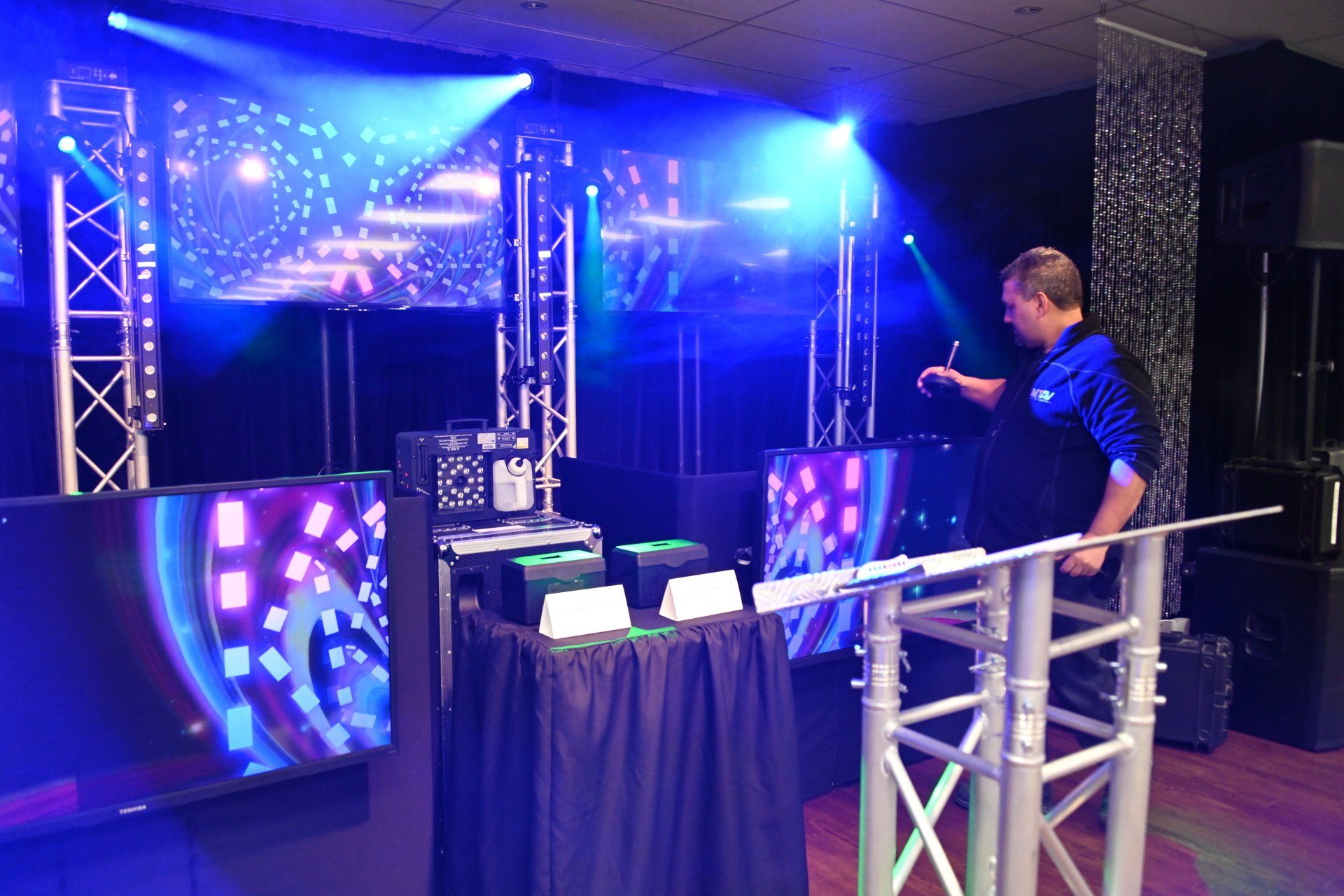 DJ setting up equipment in a room with blue stage lighting, including screens and a light rig.