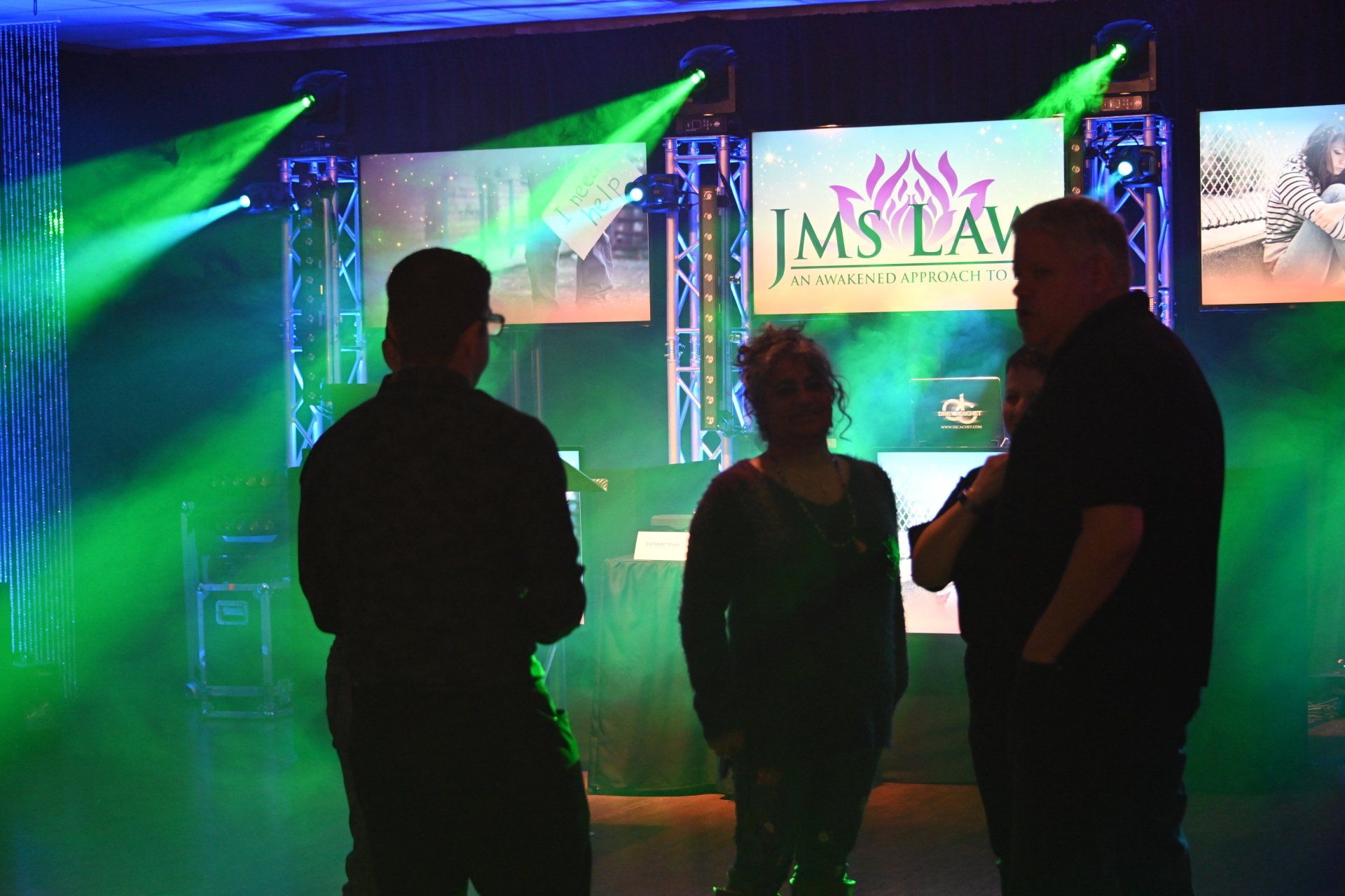 Three people in silhouette converse under stage lights and video screens displaying 