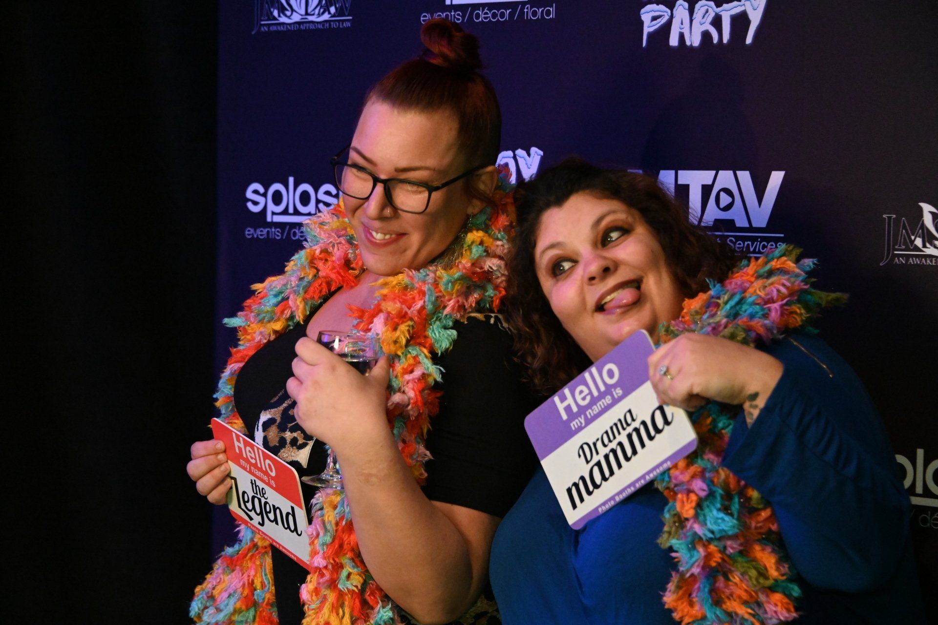 Two women at a party, holding signs and wearing leis. One smiles, the other sticks out her tongue.