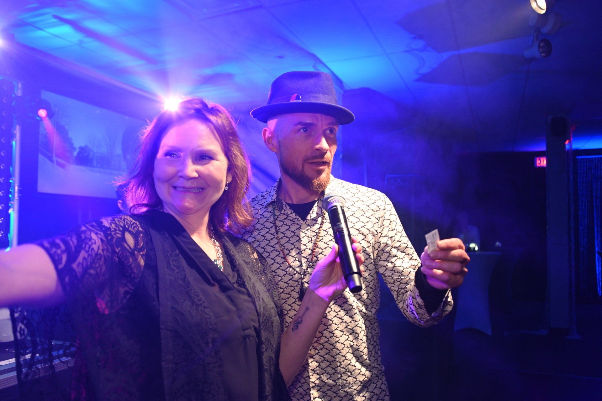 Woman and man with a microphone, in a dimly lit venue with blue and purple lighting, smiling.