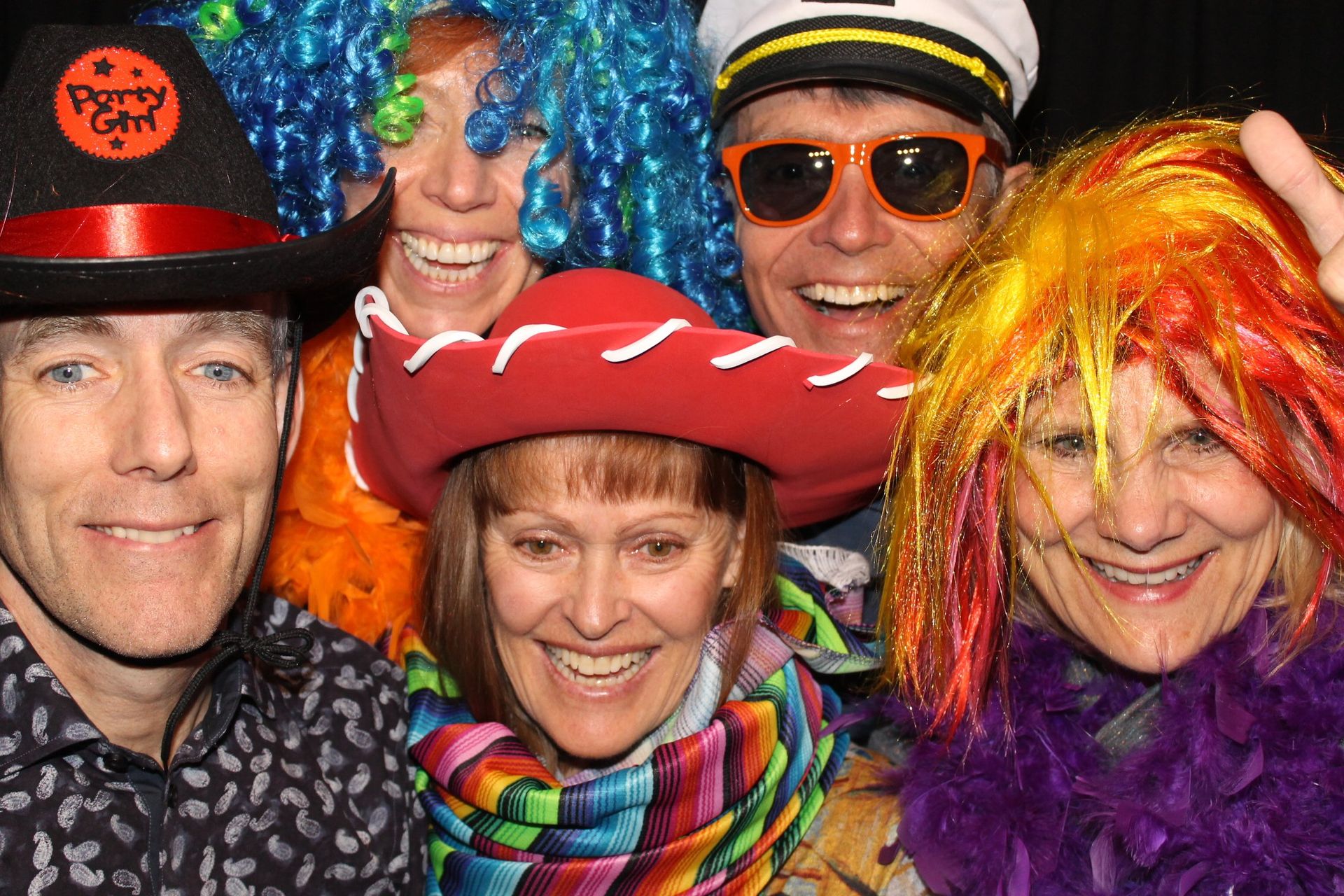 Group of people smiling, wearing colorful wigs and hats, posing for a photo at a party.