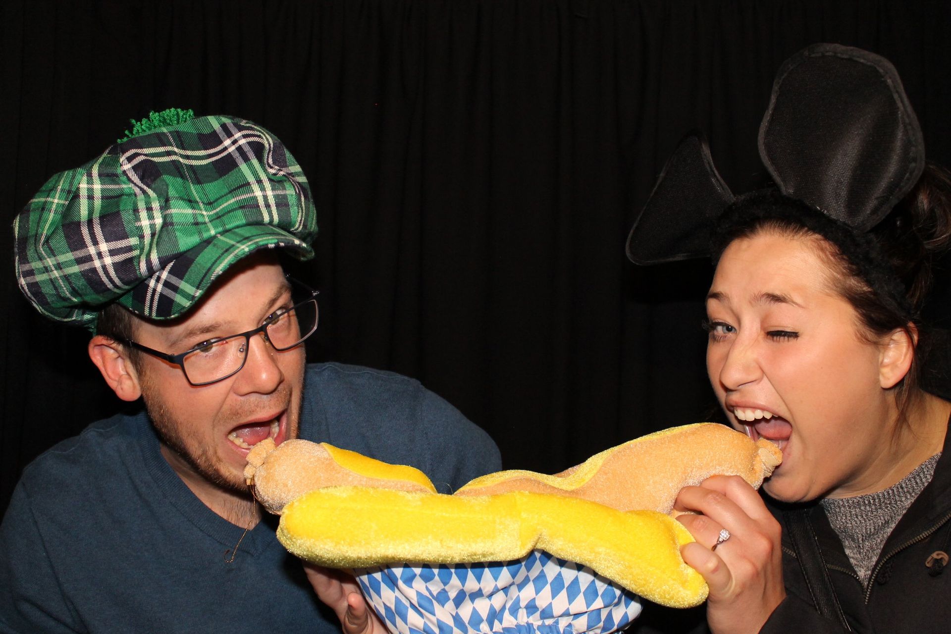 Two people, one with a plaid hat, the other with mouse ears, eating a giant yellow food item together.