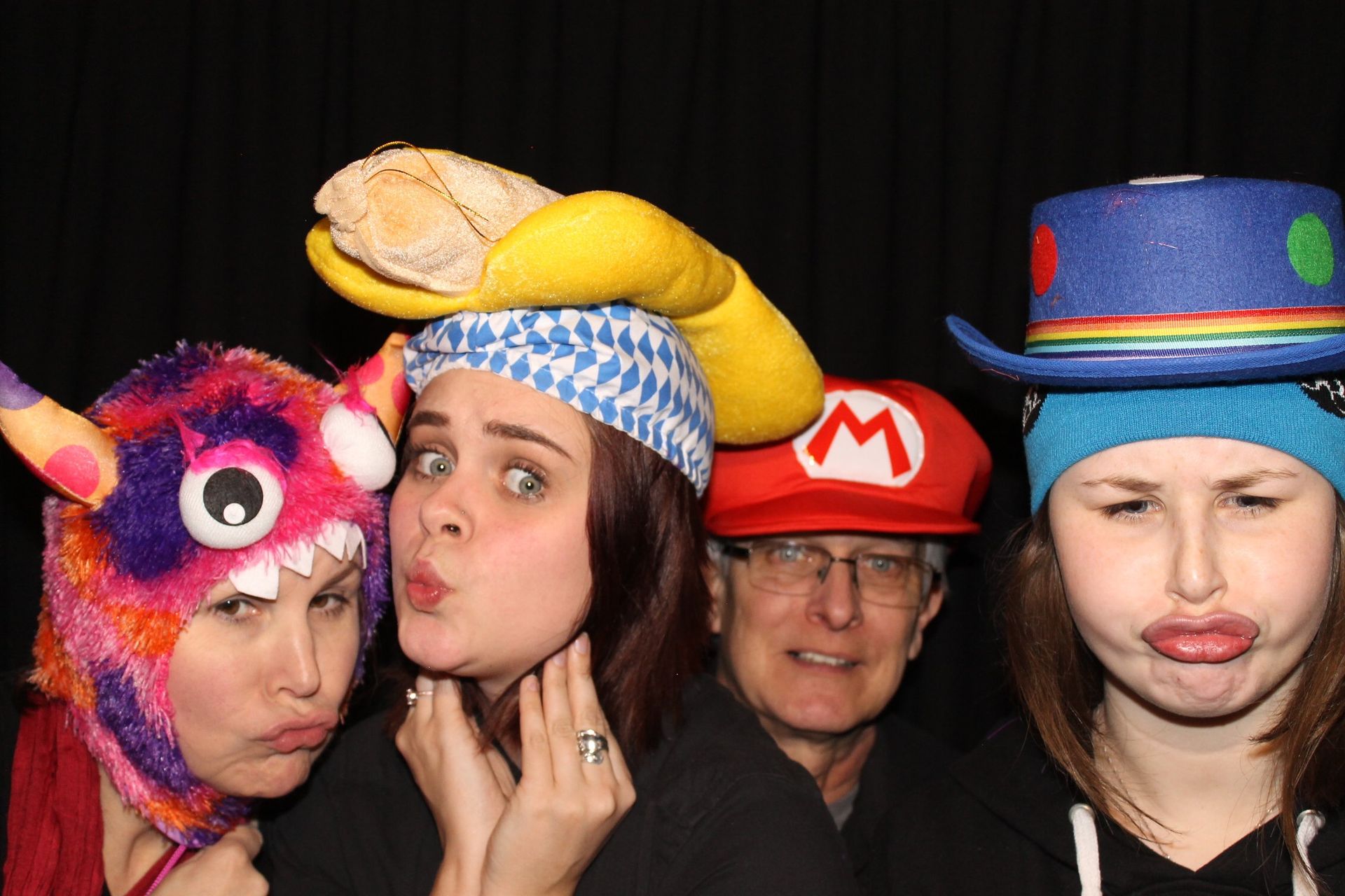 Four people in silly hats make funny faces in a photo booth, including a monster, a loaf of bread, and a Mario hat.