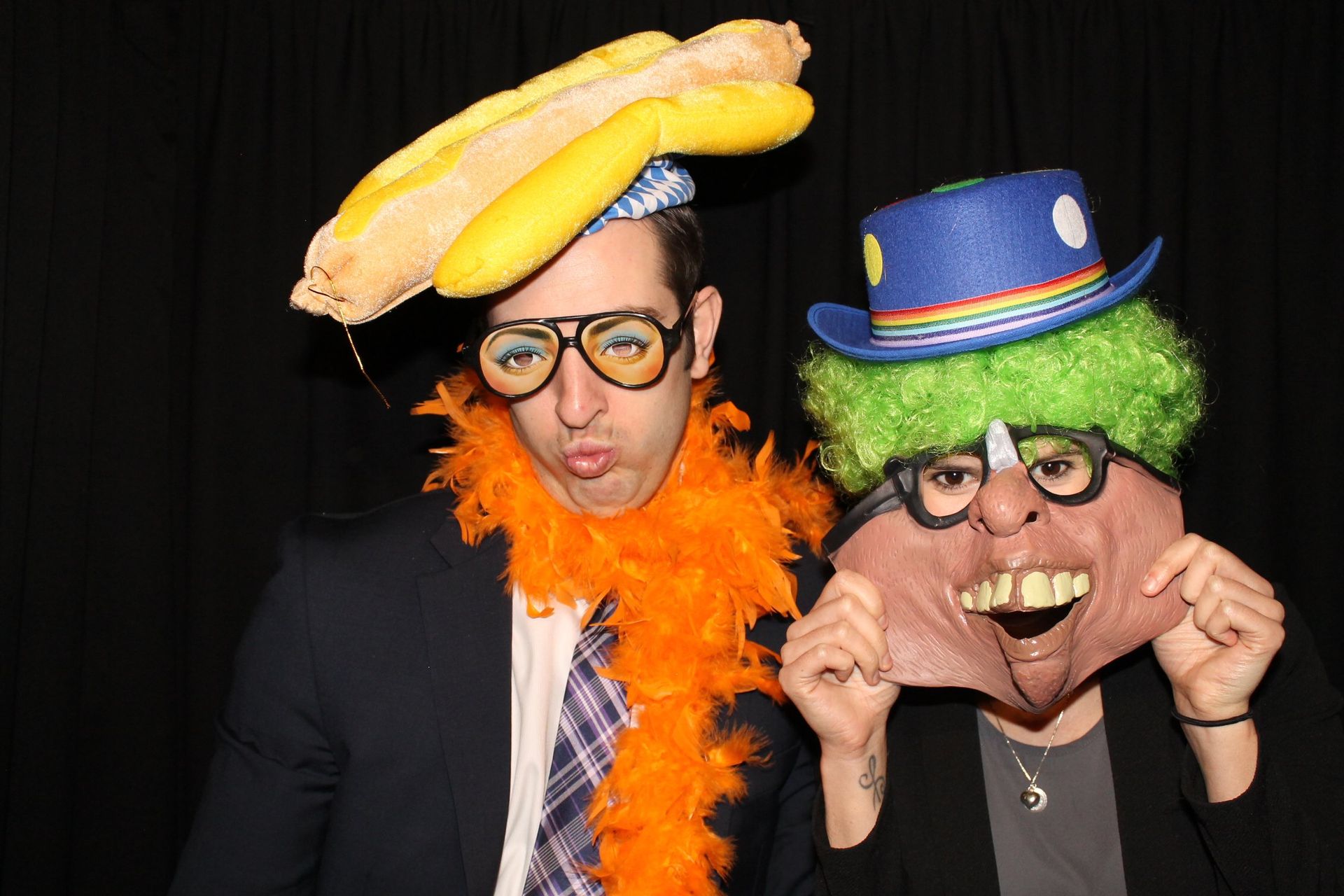 Two people wearing silly hats and accessories, making faces in front of a black backdrop.