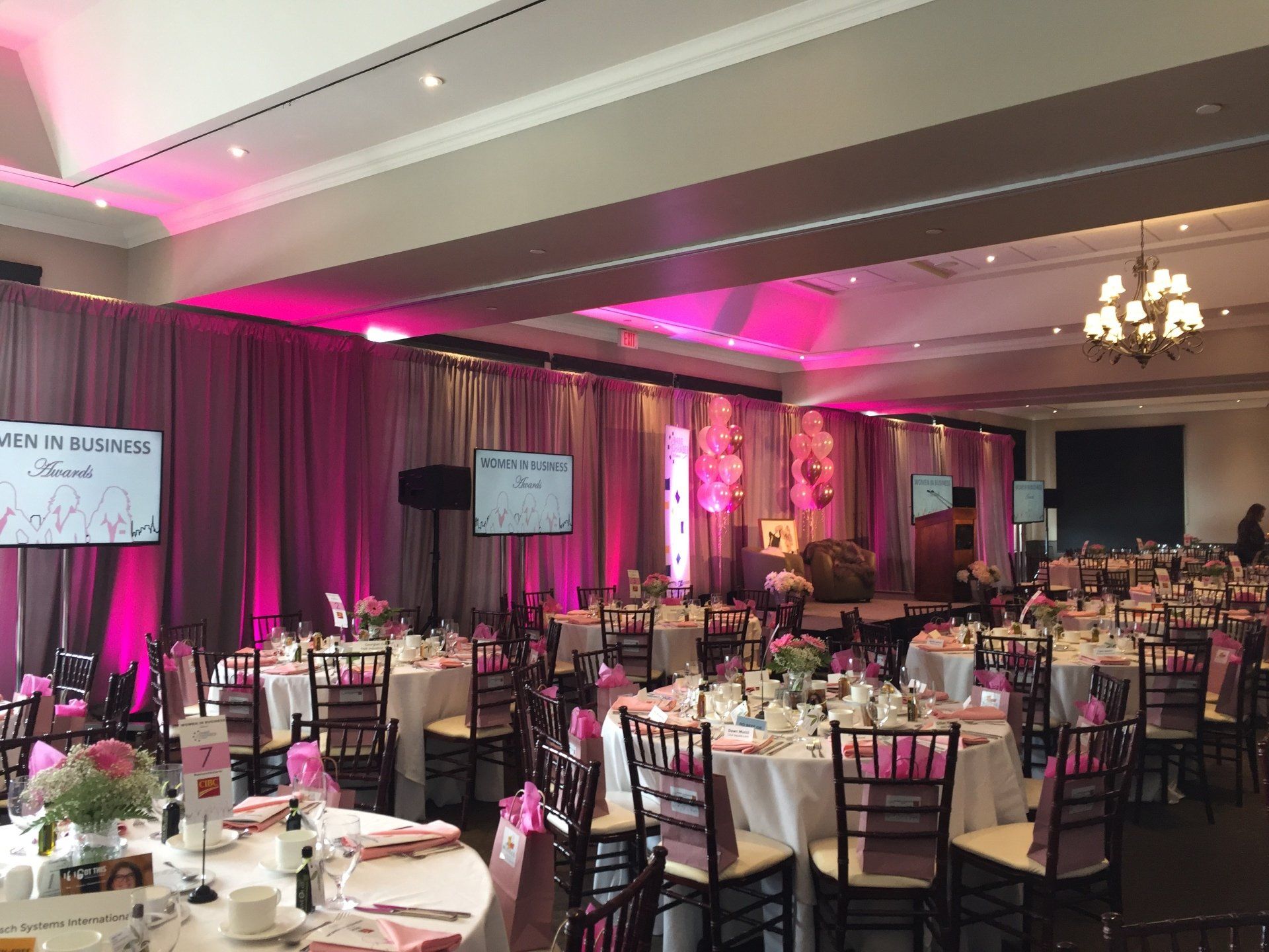 Banquet hall with round tables set for a formal event; pink uplighting, drapes, and floral centerpieces.