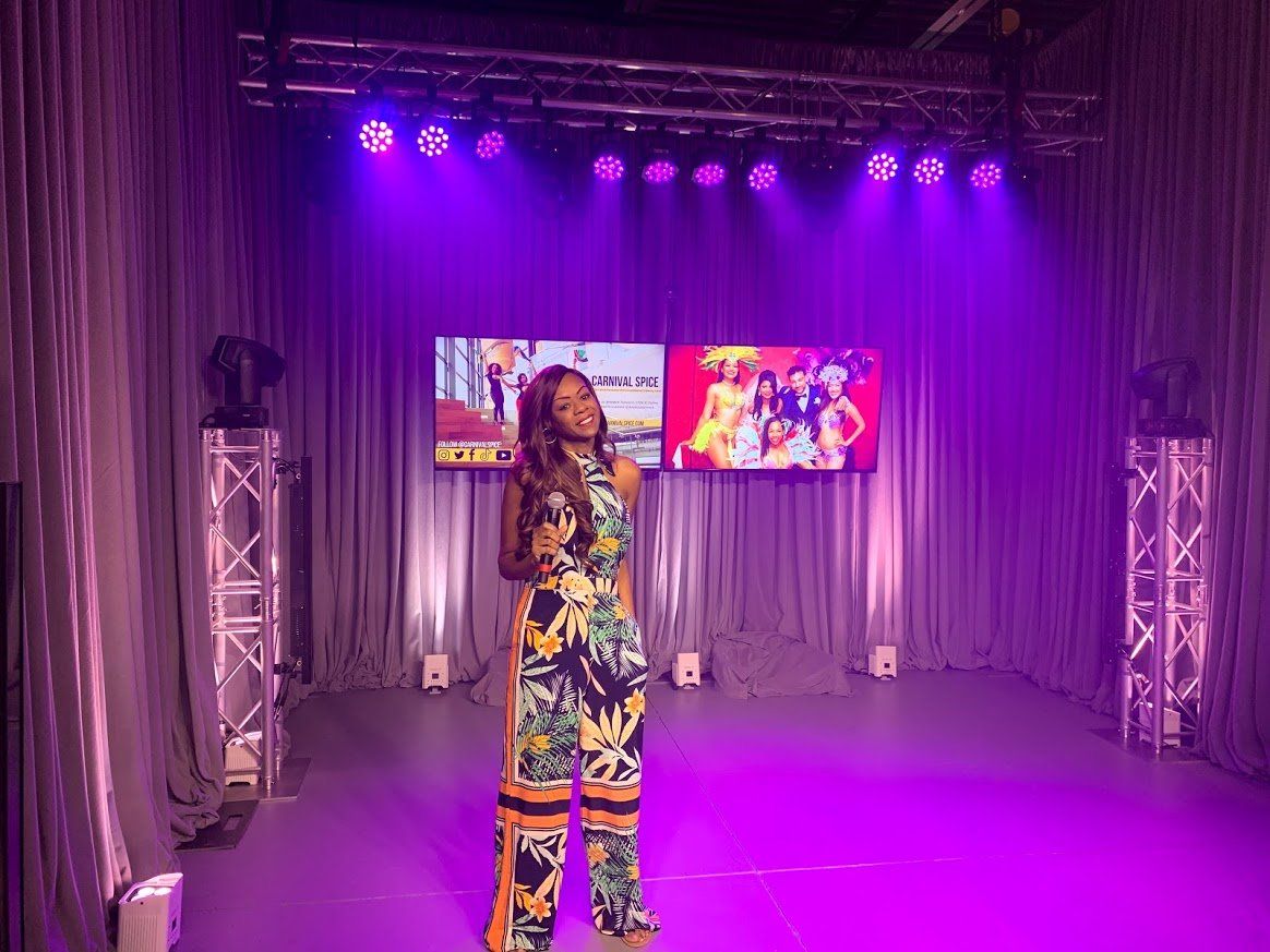 Woman in jumpsuit stands on a stage lit with purple lights, two screens in the background.