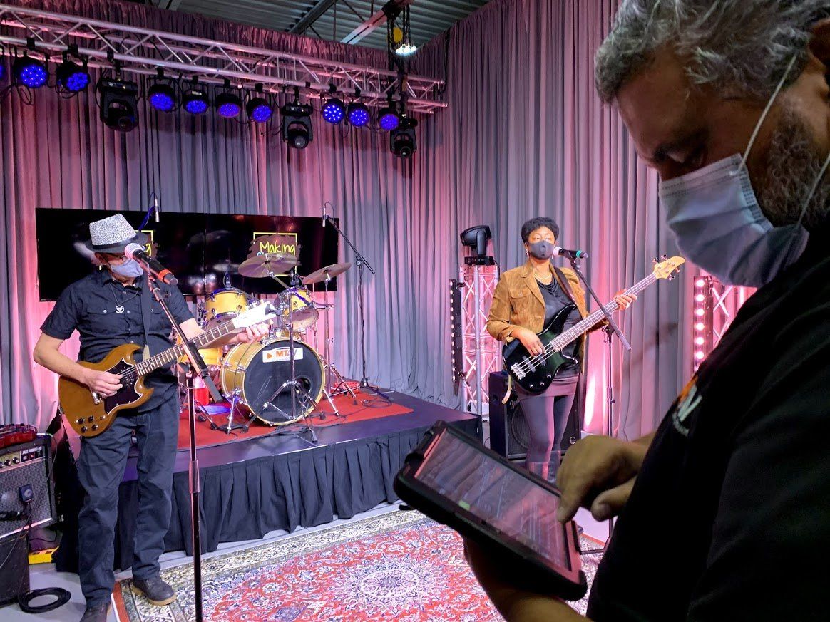 Band playing on stage with a person using a tablet in the foreground. They are wearing masks.