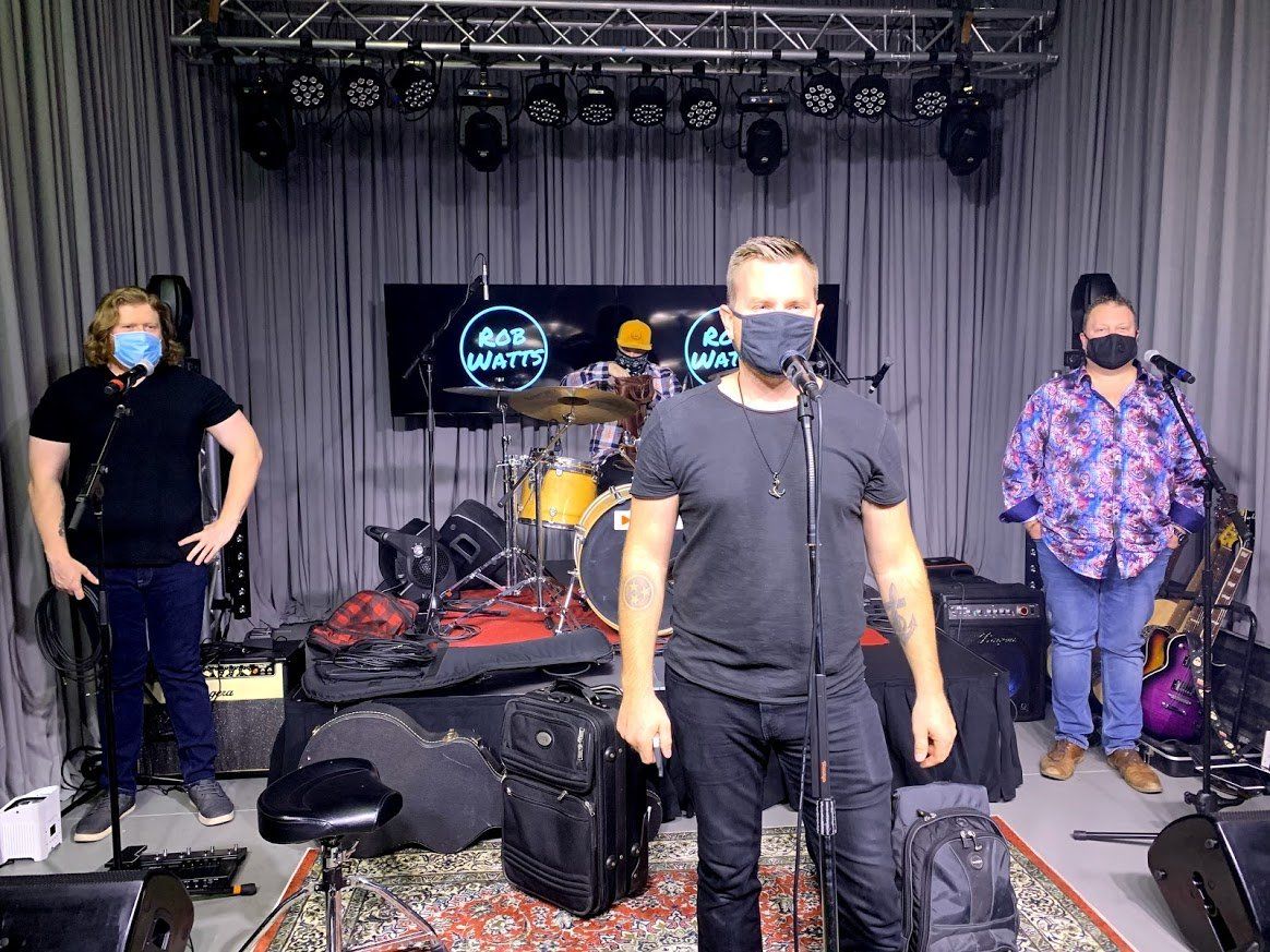 Three people in a music studio, two wearing masks, drums and equipment visible.