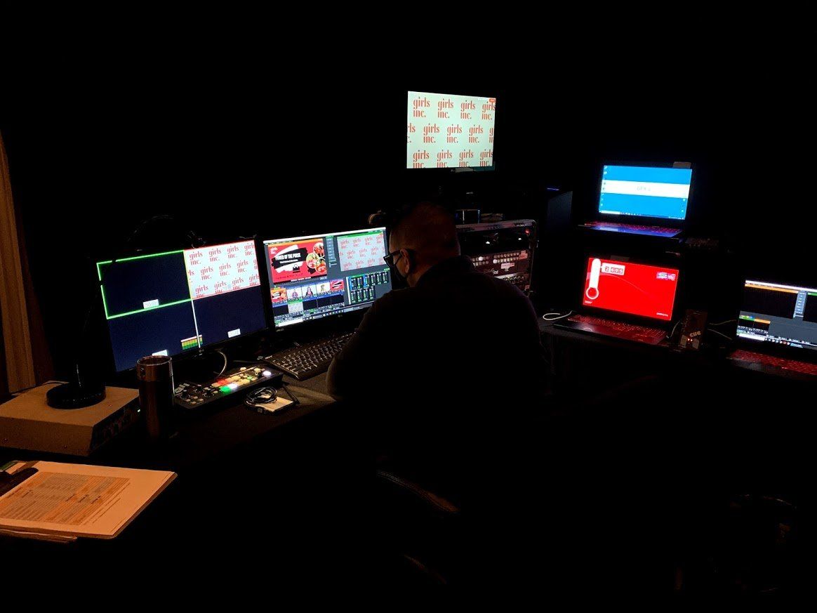 Man at a control panel in a dark room with multiple monitors displaying video feeds.