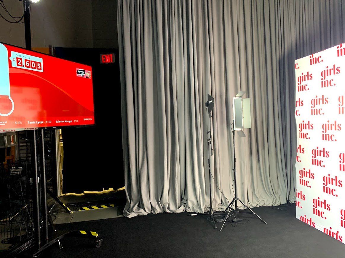 Event setup with Girls Inc. backdrop, screen, and curtains. Lighting equipment on tripods.